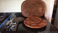 Photo showing dark stained rectangular wooden serving tray, round wooden lazy Susan, vintage cast iron waffle iron, and various stainless steel kitchen tools on black counter.