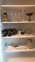 Three pantry shelves holding a variety of glassware, ceramic cups, and silver plate items including a tall beer stein, champagne glasses, mugs, and a large trophy cup.