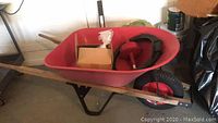 Metal wheelbarrow with red basin, wooden handles, black metal frame, and red pneumatic wheel along with box containing red tool and coiled fishtape inside basin.