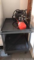 Air compressor resting on top shelf of black utility table with visible hose coiled on top