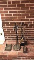 Fireplace tools set including two stands and multiple tools arranged against a brick fireplace.