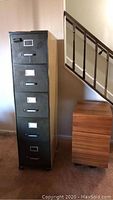 Photo showing tall metal five-drawer vertical file cabinet with black finish and smaller wooden two-drawer file cabinet beside it under a staircase.