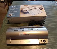 Mainstays laminator on table with its power cord and original packaging box, showing the product image and features.
