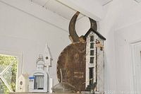 Photo shows tall narrow white bird house with multiple windows and staircase, placed beside the African wooden mask with curved horns and the clear glass wine decanter.