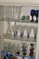 Wide shot of cabinet shelves displaying clear and colored barware including tall glasses, decanter, stemmed glasses