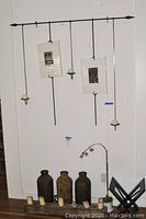 Full view of the wall hanger with hanging rods, picture frames, candle holders, ceramic vases below with candles in front, and silk plant on the floor next to a black magazines rack.