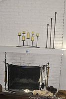 Overall view of curved fireplace screen with fireplace tool set, decorative sword, multi-arm and slim metal candle holders on white brick fireplace mantle, basket of flowers, bowl with pine cones, and burlap bag with kindling wood pieces on hearth.
