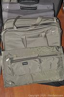 Three stacked bags: grey suitcase at back, black briefcase in middle, olive green garment bag in front