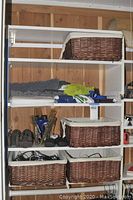 White shelving unit with four brown woven baskets lined with cream fabric. Cleaning items such as Swiffer mop heads, dusters, hand tools, and spray bottles on shelves. Gray rug runner folded on shelf.