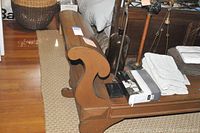 Side view showing one scrolled armrest and leg detail of the large wooden bench on a woven rug.