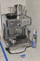 Full view of machine on tiled counter with portafilter attached, steam wand, drip tray and power cord next to soda bottle for scale