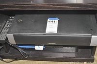 Front view of Bose sound system main unit, black body with silver vertical sides, placed on shelf with power cord coiled beside it.