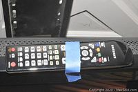 Samsung TV remote control showing various buttons and Samsung branding. Blue tape secures the remote.