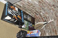 Shows packaged Rollerblade inline skates, volleyball game set, wooden paddle racket, fire extinguisher box, and black bicycle helmet on carpet against brick wall.