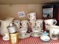 Wide view of the lot showing coffee and tea cups, cream pitcher, sugar container, vintage Lipton's Tea tin, and metal tea balls all arranged on red and white checkered fabric.