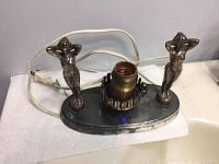 Full view of the metal table lamp base with two reclining metal figurines and power cord.