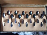 Twelve ornate silver teaspoons arranged inside a tan fabric-lined presentation case with hinged lid.