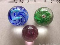 Three glass paperweights on a table with a lace tablecloth, showing different angles of the blue dolphin swirl, green spiral floral, and purple swirl designs.