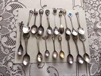 Photo showing all 15 souvenir spoons laid out, displaying varied handle decorations including enamel crests, architectural likenesses, and metallic relief work