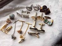 Photo showing various pairs of men's cufflinks with different designs including gold-tone ovals, silver rectangulars, turquoise inlays, and geometric shapes, as well as tie clips with chain attachments.