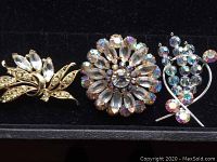 Three vintage brooches displayed on black velvet: gold-tone leaf brooch with clear rhinestones, circular floral brooch with clear and aurora borealis rhinestones, silver-tone floral spray brooch with aurora borealis rhinestones.