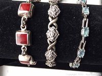 Photograph showing close-up view of three sterling silver bracelets displayed on black background. Red square stones, textured silver design, and blue oval stones are visible.