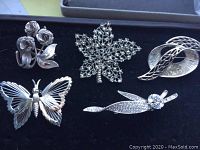 Five silvertone brooches arranged in black tray: rose flower, maple leaf with rhinestones, butterfly with wire detail, abstract swirl, leaf with rhinestones.