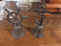 Two cast iron nautical bells on wooden surface, showing front view of ship wheel and anchor designs
