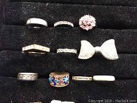 Rings arranged on black holder show various costume rings including floral, rhinestone bands, enamel work, and bow-shaped ring.