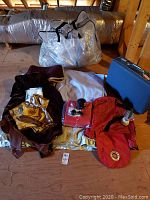 Photo showing assortment of blankets, throws, satin quilt, plastic zip storage bags, vintage suitcase and some red waterproof materials with lantern and other small items on wooden floor in attic.
