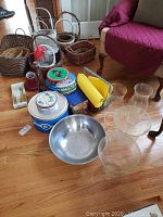 Photo showing 12 assorted woven baskets, six decorative tins, and a large metal mixing bowl on floor with glass vases and lamp shades in background.