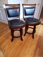 Pair of cherry wood swivel bar chairs with black vinyl upholstery and visible wear on vinyl surfaces