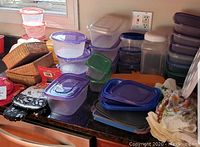 Wide view showing over 28 plastic food storage containers stacked and grouped, mostly rectangular and square shapes with blue, purple, and green lids. Several kitchen textiles including pot holders and oven mitts to the side. Wicker baskets and miscellaneous items at edge of counter.