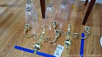 Two brass wall sconces side by side with four clear glass chimneys placed behind them on the floor. Blue tape markers on floor for sizing.