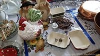 Photo shows chicken cookie jar front with visible green and red painted details, snowman plush figures behind, plus white and red stoneware trays on table.
