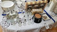 Shows multiple sets of glass and crystal salt and pepper shakers, three 4-packs of Bombay Company place card holders, a set of Bombay place card holder frames, cork and wood coasters, and assorted wine glass charms on a white tablecloth.