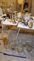 Two white tables with silverplated candle holders, napkin rings, trinket boxes, and glass vases on a basement floor.