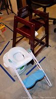 Full view of the wooden WinCo high chair along with the white and turquoise plastic step-up toilet trainer and toddler potty training seat, placed on floor marking taped lines.