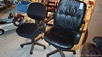 Two office chairs side by side on a concrete floor. One black high-back vinyl chair showing wear, next to a black fabric mid-back swivel chair with armrests.