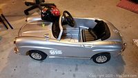 Silver Mercedes Benz child's electric ride-on car with black seat and steering wheel, including a red and black child's helmet placed on the car's back seat area