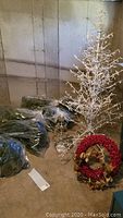 Overall view showing 6ft white metal tree with white string lights, two wreaths (one red with pine cones), and bags likely containing multiple small battery-operated outdoor trees.