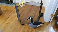 Photo showing the wooden decorative goose stand placed in front of the brass fireplace screen on wood flooring.