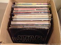 Box of over 60 LP records stacked upright showing a few recognizable album covers including Star Wars soundtrack at the front.