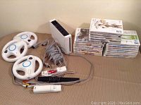 Photo showing Nintendo Wii console, two white steering wheel controllers, sensor bar, Wii remote missing the back cover, AV cables, power adapter, and stacked Wii game cases.