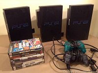 Three PlayStation 2 consoles, stacked vertically, with four PlayStation 2 controller cables coiled and eight PS2 game cases stacked beside consoles.