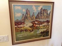 Framed oil painting showing mountain houses by a water body with mountainous peaks in the background and blue cloudy sky.