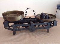 Full view of antique balance scale with black metal frame and two brass pans, one side holds four weights.