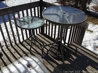 Outdoor photo showing bar height patio table with textured glass top next to smaller round side table with glass top on a wooden deck with snow patches.
