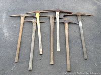Seven garden picks laid out on pavement showing wood handles and rusted metal heads, some handles loose or worn.