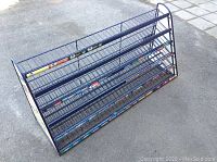 Front and side diagonal view of a blue metal wire candy display rack with four shelves, placed on pavement.